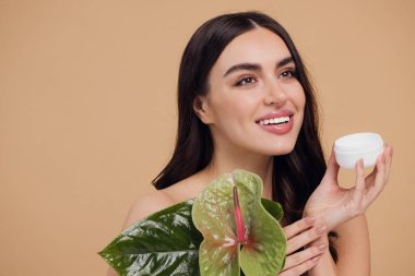 Yeşil çiçeğin yanındaki çekici bir kadının portresi sorun için yüz kremi sürüyor. Bej arka planda merhem var. Refah konsepti