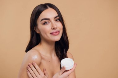 Gülen, yarı çıplak, gülümseyen ve bej arka planda izole edilmiş yüz kremi süren güzel bir kadının güzel portresini kapat.