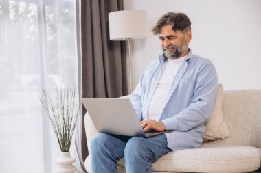Oturma odasında dizüstü bilgisayar kullanarak gülümseyen olgun bir adam. Mutlu kendinden emin yetişkin erkek evde dinleniyor bilgisayar üzerinde çalışıyor, klavyede daktilo yazıyor, boşluğu kopyalıyor
