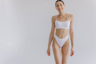 İnce vücutlu güzel bir kadın ve beyaz iç çamaşırlarıyla beyaz stüdyo arka planında poz veren mükemmel bir midesi var..