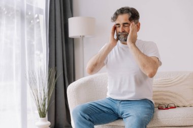 Home, senior man and headache on sofa for stress, emotional conflict and temporal vertigo. Living room, couch and old person with pain, migraine and anxiety due overthinking in retirement in house clipart