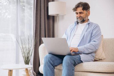 Oturma odasında dizüstü bilgisayar kullanarak gülümseyen olgun bir adam. Mutlu kendinden emin yetişkin erkek evde dinleniyor bilgisayar üzerinde çalışıyor, klavyede daktilo yazıyor, boşluğu kopyalıyor