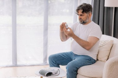 Oturma odasındaki koltukta oturan yaşlı adam kan basıncını cihazla ölçüyor. Yaşlı erkek hapları tutuyor ve uyuşturucu bırakıyor. Sağlık ve öz bakım kavramı, kopyalama alanı