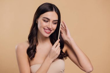 Çıplak omuzlarını bayanla su kuyusu-damat rüya gibi esmer saç o ahşap fırça pastel bej renkli arka plan üzerinde izole ile saç tarama büyüleyici portre yapmak beyaz ışınlama dişleri kazma dişli bir gülümseme