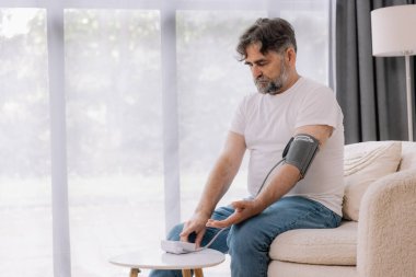 Evdeki kanepede oturmuş, tansiyonunu ölçen, gri saçlı, son sınıf öğrencisi. Modern tonometre kullanan emekli, hipertansiyon deneyimi yaşayan, kopyalama alanı