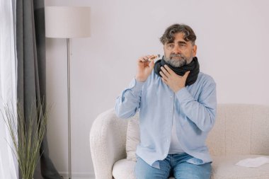 Yüksek ateşi olan yaşlı bir adam, evdeyken termometresini dikkatlice kontrol ediyor. Etrafı çeşitli ilaçlarla çevrili bir battaniyeye sarılmış ve kendini pek iyi hissetmiyor ve endişeli görünüyor..