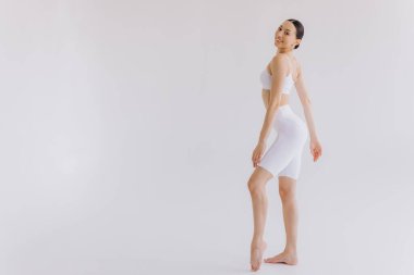 Beyaz arka planda beyaz spor giysili zayıf bir kadın var. Düz göbekli, kaslı, zayıf, çekici bir kadın. Uzayı metin için kopyala. Vücut bakımı, sağlıklı ve sportif yaşam, saç aldırma, yoga konsepti