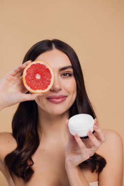 Kadın, doğal cilt bakımı için nemlendirici krem ve greyfurt ve bej renkli stüdyo arka planına karşı C vitamini. Sağlıklı organik ürünler için meyve ve krem ya da losyon tutan mutlu kadın portresi