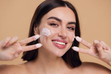 Kadın güzel, mutlu yüz, krem sürerek ve cildini temizlerken parmakları, tırnağı ve saçı, güzel saçı, vücudu ve bej arka planda güzellik konsepti.