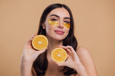 Güzel, saf, kibar bir kadının çıplak, izole bir şekilde duran görüntüsü. Gözlerinin altında altın bantlarla cildine dikkat et. Bej arka planda yarım portakal ile fotokopi alanı tut.