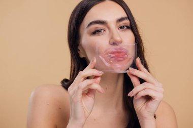 Beautiful Happy Woman holding collagen lip mask for dry and chapped lips on Beige Background. Beauty concept portrait. Pink hydrogel lips patch. Korean cosmetics. clipart