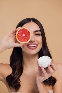 Kadın, doğal cilt bakımı için nemlendirici krem ve greyfurt ve bej renkli stüdyo arka planına karşı C vitamini. Sağlıklı organik ürünler için meyve ve krem ya da losyon tutan mutlu kadın portresi