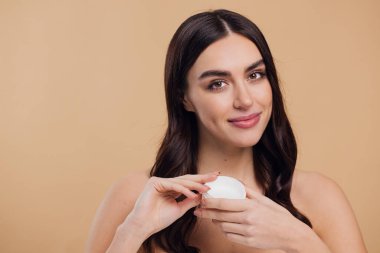 Yarı çıplak, mutlu, mutlu, sağlıklı bir cilde sahip bir kadının bej arka planda izole edilmiş yüz kremi sürüşü.