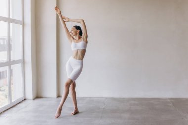 Zayıf vücutlu sportif genç bir kadın, panoramik pencereli modern odada yoga pratiği yapıyor. Vücut ve zihinsel gelişim arasındaki sağlıklı yaşam ve doğal denge kavramı.