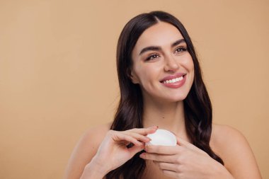 Yarı çıplak, mutlu, mutlu, sağlıklı bir cilde sahip bir kadının bej arka planda izole edilmiş yüz kremi sürüşü.