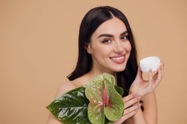 Yeşil çiçeğin yanındaki çekici bir kadının portresi sorun için yüz kremi sürüyor. Bej arka planda merhem var. Refah konsepti