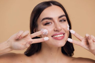 Kadın güzel, mutlu yüz, krem sürerek ve cildini temizlerken parmakları, tırnağı ve saçı, güzel saçı, vücudu ve bej arka planda güzellik konsepti.