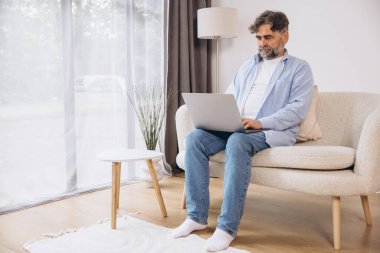 Oturma odasında dizüstü bilgisayar kullanarak gülümseyen olgun bir adam. Mutlu kendinden emin yetişkin erkek evde dinleniyor bilgisayar üzerinde çalışıyor, klavyede daktilo yazıyor, vücut uzunluğu, kopyalama alanı