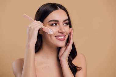 Yarı çıplak kadın, güzellik için yüz ve fırça kremi, kuaför ve cilt bakımı bej arka planda. Mutlu model, yüz losyonu ve dermatoloji, kolajen ve mutluluğun gülümsemesi için makyaj malzemesi.