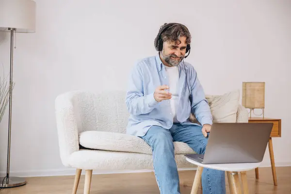 stock image Positive grandfather using laptop computer and headset, sitting on couch. Senior man working from home, have online meeting, video chat, copy space