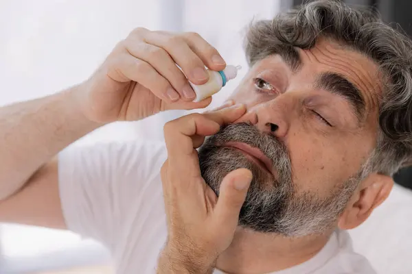 stock image Close-up photo of an older gray-faced man who drips eye drops with medicine for pain and fatigue, sitting on the couch at home.