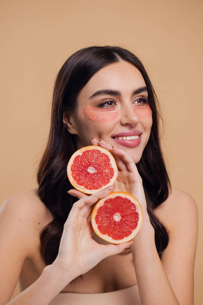Beauty Image of young pure beautiful gentle woman standing naked isolated looking camera take care of her skin with under eye pink patches and holding half grapefruits on beige background with copy space