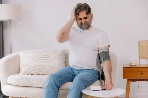 stock image Handsome grey-haired senior man in casual outfit sitting on couch at home, checking blood pressure on his own. Pensioner using modern tonometer, experiencing hypertension, copy space