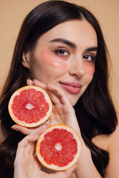 Beauty Image of young pure beautiful gentle woman standing naked isolated looking camera take care of her skin with under eye pink patches and holding half grapefruits on beige background with copy space