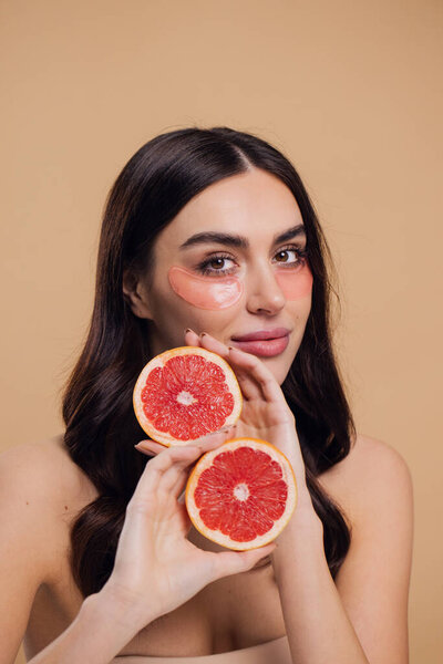 Beauty Image of young pure beautiful gentle woman standing naked isolated looking camera take care of her skin with under eye pink patches and holding half grapefruits on beige background with copy space