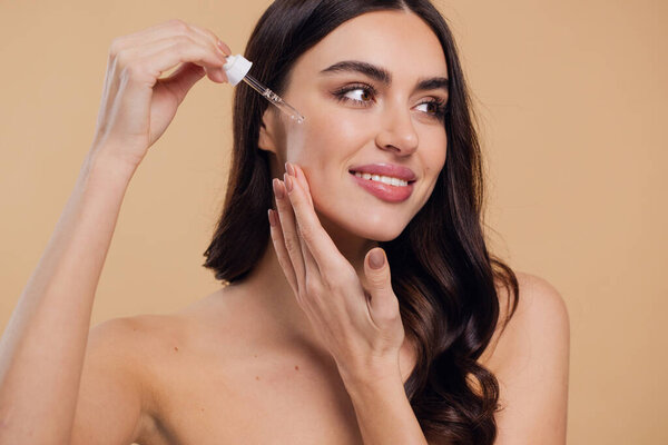 Woman Dropping Serum on Face on Beige Background. European Girl Applying Serum Collagen Moisturizer from Pipette on Face close up. Positive Laughing Beauty Woman Portrait. Concept of Face Skin Care