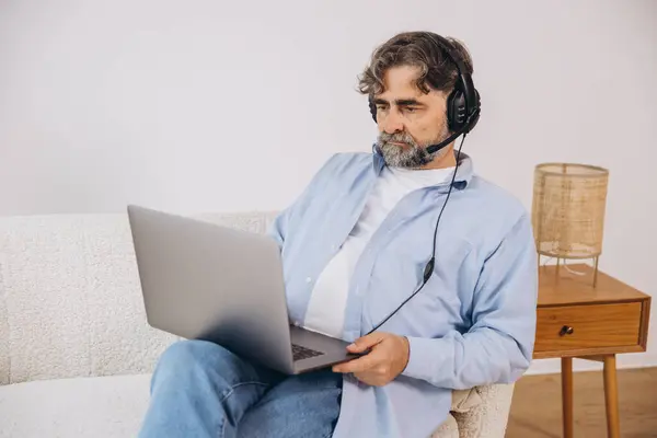 stock image Positive grandfather using laptop computer and headset, sitting on couch. Senior man working from home, have online meeting, video chat, copy space