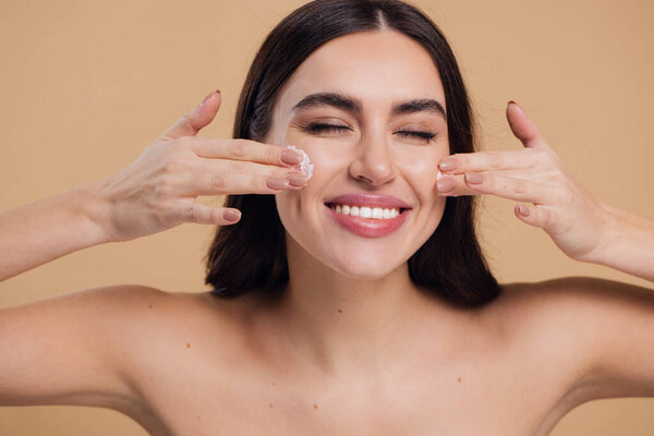 Woman beauty happy face close-up applying cream and cleanse skin with fingers of her hand, nail and hair health, beautiful hair, body and beauty care concept on beige background