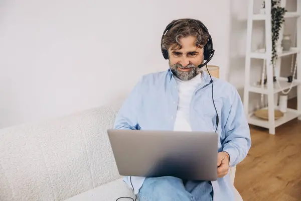 stock image Positive grandfather using laptop computer and headset, sitting on couch. Senior man working from home, have online meeting, video chat, copy space