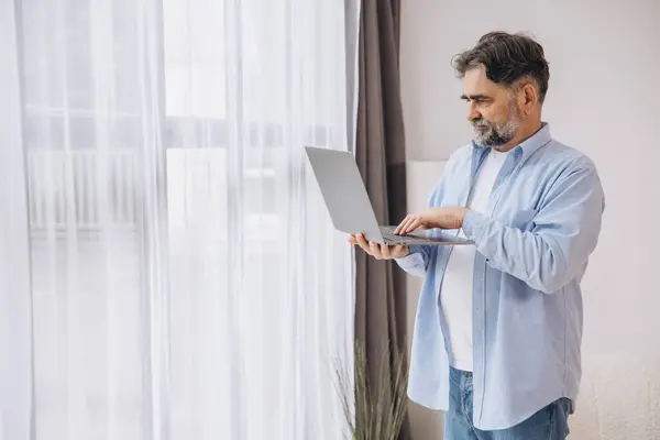 stock image Smiling mature man using laptop in living room. Happy confident senior male adult resting at home working on pc, typing on keyboard, copy space