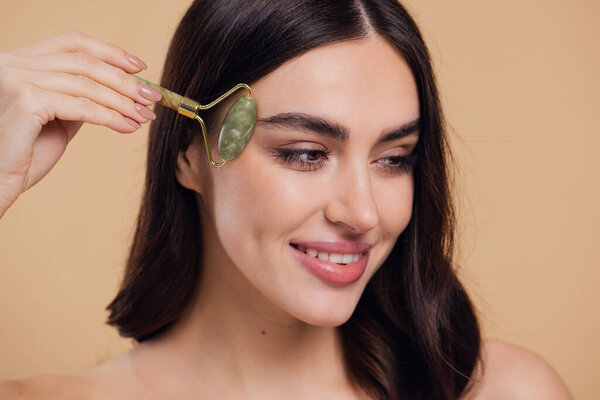 Закрыть полуобнаженный портрет красивой молодой женщины с идеальным кожным массажем лица с помощью нефритового ролика с натуральным кварцевым камнем на бежевом фоне.