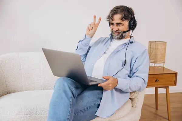 stock image Positive grandfather using laptop computer and headset, sitting on couch. Senior man working from home, have online meeting, video chat, copy space