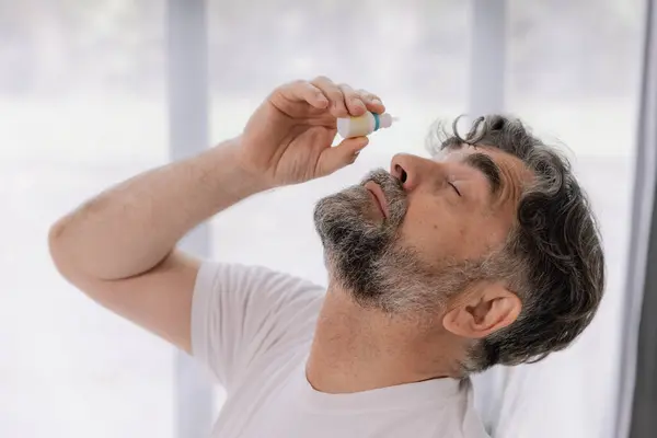 stock image Close-up photo of an older gray-faced man who drips eye drops with medicine for pain and fatigue, sitting on the couch at home.