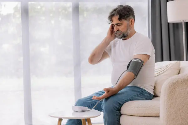 stock image Handsome grey-haired senior man in casual outfit sitting on couch at home, checking blood pressure on his own. Pensioner using modern tonometer, experiencing hypertension, copy space