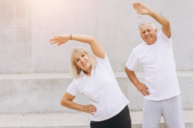 Üst düzey çift dışarıda egzersiz yapmayı seviyor. Birlikte esneme egzersizleri yapıyorlar, fitness ve sağlıklı yaşamı destekliyorlar. Her ikisi de gülümsüyor ve enerjik görünüyorlar, aktif yaşlanma faydalarını vurguluyorlar..
