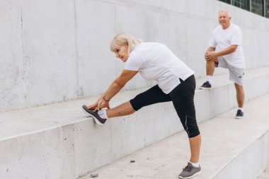 Dışarıda esneme egzersizi yapan yaşlı bir çift. Kadın ayağını beton bir çıkıntıya uzatırken erkek arka planda geriniyor. İkisi de spor kıyafetleri giymiş..