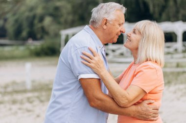 Kıdemli çift dışarıda kucaklaşıyor ve gülümsüyor, bir park ortamında mutluluk ve aşkın tadını çıkarıyorlar..