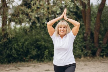 Parkta yoga yapmaktan hoşlanan yaşlı bir kadın gülümseyerek aktif ve sağlıklı bir yaşam tarzı sergiliyor..