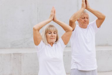 Üst düzey bir çift dışarıda beyaz giysiler içinde yoga yapıyor, sağlık ve sağlığa odaklanıyor. Yaşlı çift, elleri havada yoga pozu vererek sağlıklı ve aktif bir yaşam tarzını destekliyor..