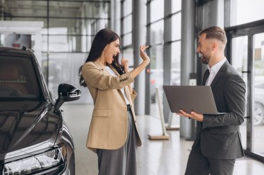 Yeni arabanın anahtarlarını tutan, yumruğunu gösterip modern araba galerisinde evet diyen beyaz bir kadın. Mutlu alıcı kız rüyasını gerçekleştirdikten sonra sevinir..