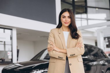 Araba Bayilik ziyaret etti. Güzel genç kadın kameraya bakarak ve çapraz kolları olan dururken yeni arabasını gülümseyen