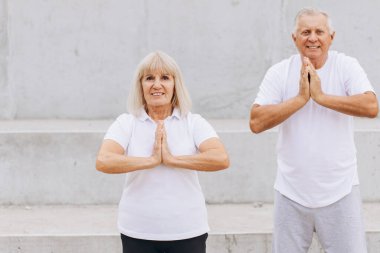 Yaşlı bir çift dışarıda yoga egzersizleri yapıyor, spor, sağlık ve rahatlama konularını vurguluyor. Dışarıdaki yoga çalışmaları sırasında beyaz atletizm kıyafetleri giyiyorlar, huzur, denge ve sağlık yayıyorlar..