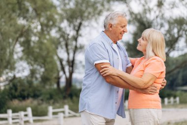 Kıdemli çift parkta gülümsüyor ve kucaklaşıyorlar. Sevgi, mutluluk ve birlikteliği doğal bir ortamda gösterir.
