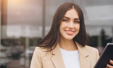 Takım elbiseli başarılı bir satıcının yakın plan portresi elinde dijital tabletle açık hava araba galerisinde duruyor.