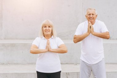 Yaşlı bir çift dışarıda yoga egzersizleri yapıyor, spor, sağlık ve rahatlama konularını vurguluyor. Dışarıdaki yoga çalışmaları sırasında beyaz atletizm kıyafetleri giyiyorlar, huzur, denge ve sağlık yayıyorlar..
