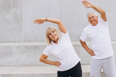 Üst düzey çift dışarıda egzersiz yapmayı seviyor. Birlikte esneme egzersizleri yapıyorlar, fitness ve sağlıklı yaşamı destekliyorlar. Her ikisi de gülümsüyor ve enerjik görünüyorlar, aktif yaşlanma faydalarını vurguluyorlar..